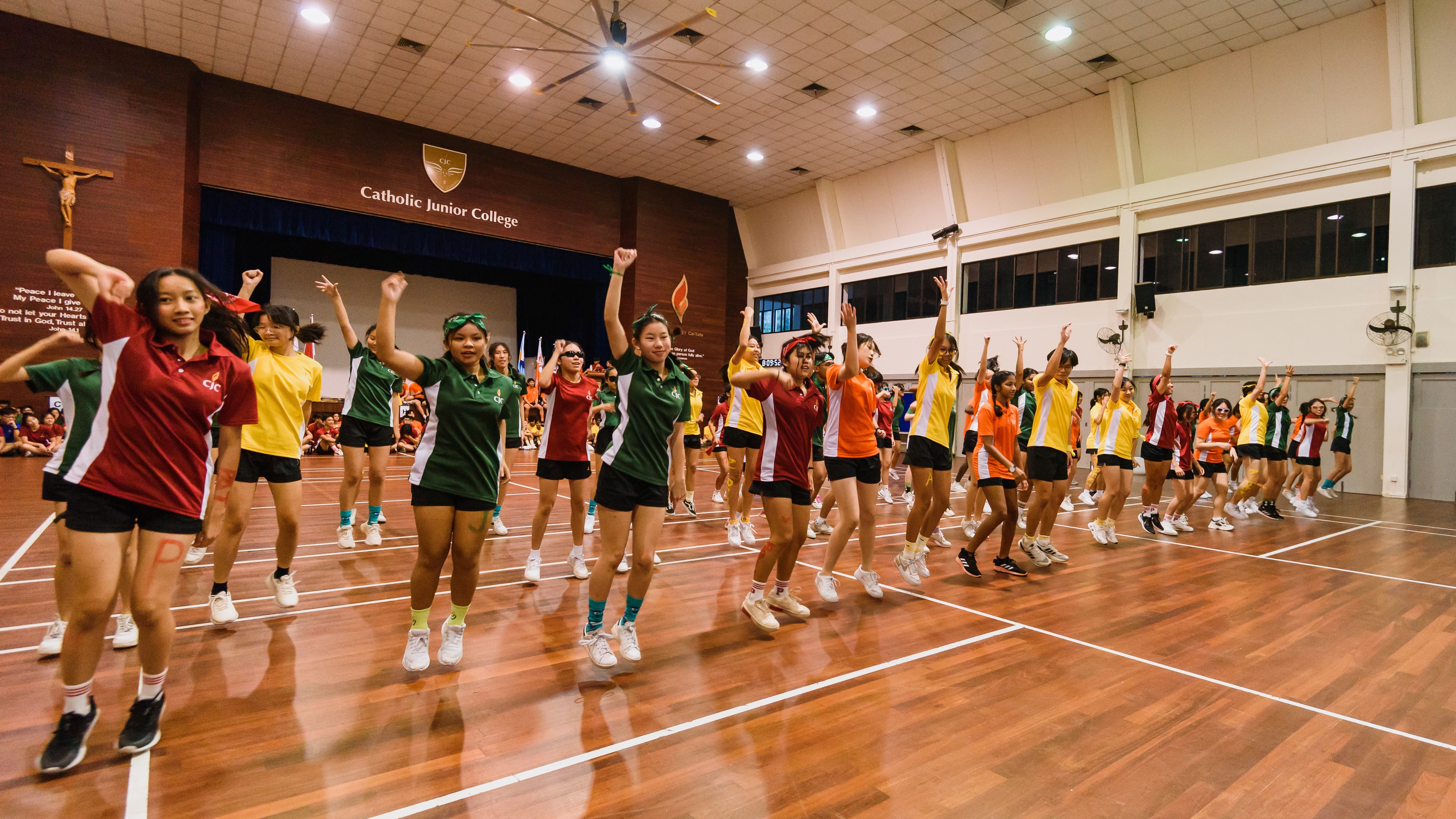 Students in colorful jerseys jump with raised arms on an indoor court under a Catholic Junior College sign.